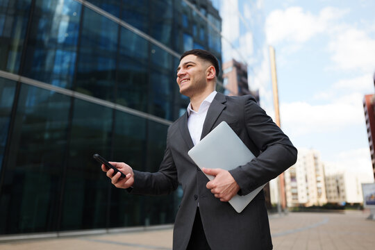 Young professional walking near buildings holding a laptop and phone