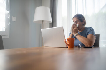 Woman working from home at dining table on laptop blowing nose with cold.