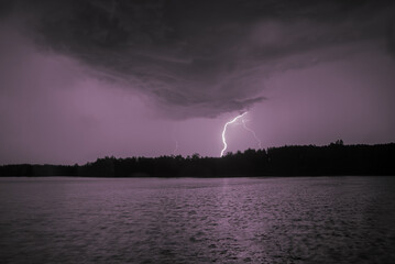 \Lightning strikes over a dark sky above a lake, illuminating clouds and water with a purple glow.