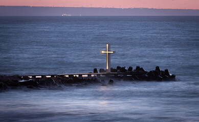 A glowing, modern cross standing on a pier at the edge of a turbulent sea. The cross, a dark silhouette with a bright, fiery orange and yellow core, serves as a beacon against the dramatic, cloudy sky