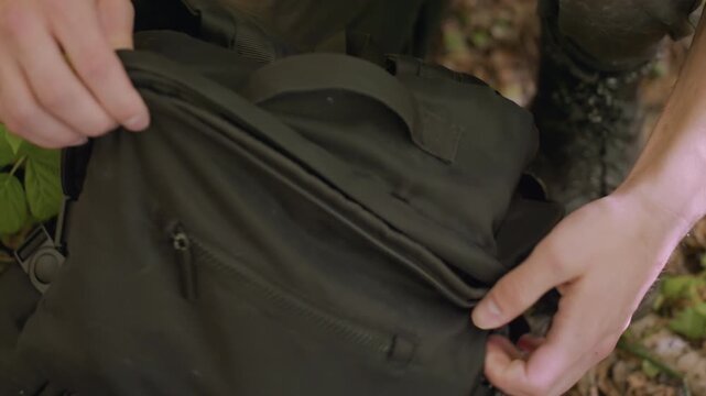 Young man unzips bag in forest, hands pulling zipper open as green leaves brush against bag, revealing blurry view of contents including gear, dirt-covered trousers, and natural surroundings