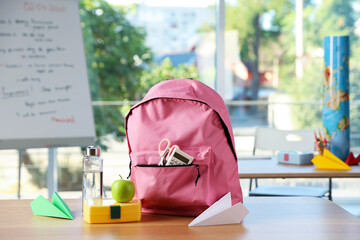 Backpack with different stationery, lunchbox, apple, bottle of water and paper planes on desk in classroom