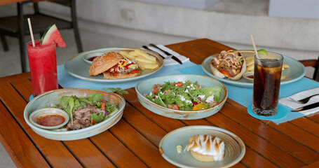 Cafe table setting with gourmet burgers, fries, fresh salads, watermelon juice, cold drinks. Vibrant, delicious dining in cozy cafe atmosphere.
