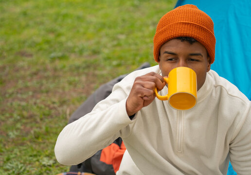 Portrait of young african american man wearing orange beanie and white fleece enjoying a hot beverage in a yellow mug while camping outdoors