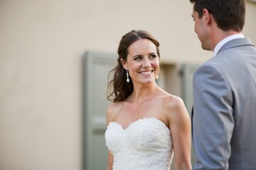 Naklejka premium A white woman in a simple wedding dress stands next to a man in a suit, both smiling softly. The background is plain with soft natural light and no extra objects