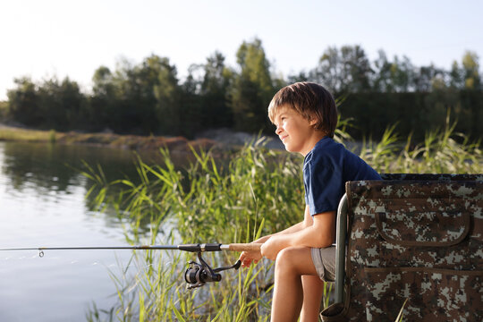Little boy sitting on folding chair and fishing near river outdoors