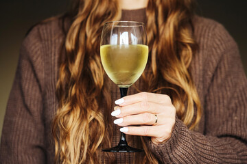 Glass of white wine in woman hand. Closeup of drink in elegant glass. Woman with long hair in sweater holding wine. Alcohol lifestyle concept. Evening relaxation moment. Cozy home atmosphere scene.