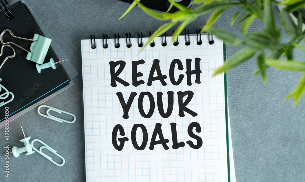 Poster a notebook with the word reach your goals written on it. the notebook is on a table with a pair of s