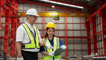 Two industrial workers wearing safety helmets and reflective vests discussing checklist inspection in a modern metal sheet factory, focusing on teamwork, safety, and quality control in production.