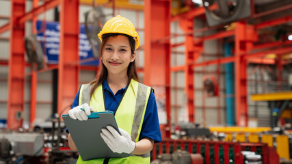 Smiling female engineer wearing safety helmet and vest holding clipboard in modern factory, representing industry, engineering, teamwork, occupational safety, and professional success.