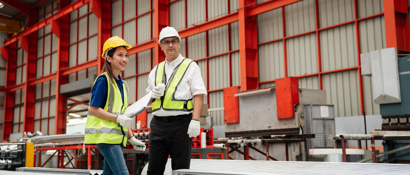 Two industrial workers wearing safety helmets and reflective vests discussing checklist inspection in a modern metal sheet factory, focusing on teamwork, safety, and quality control in production.