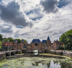 A picturesque medieval gate of the city of Amersfoort with pointed towers stands over a calm canal. The sky is filled with dramatic clouds, adding to the historic ambiance.