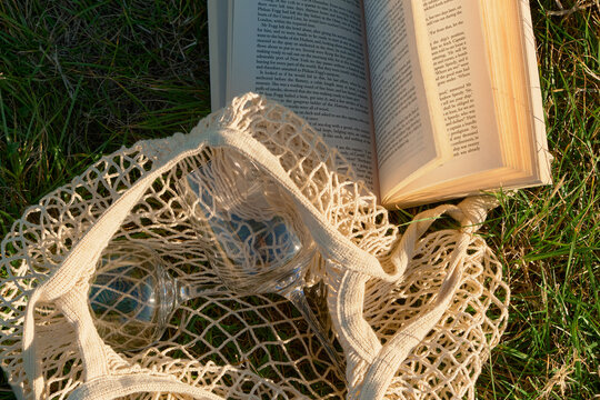 Horizontal photo highlighting the woven texture of a vintage mesh bag with wine glasses, an open book, and autumn grass. Sunlight enhances transparency of glasses and flying book pages.