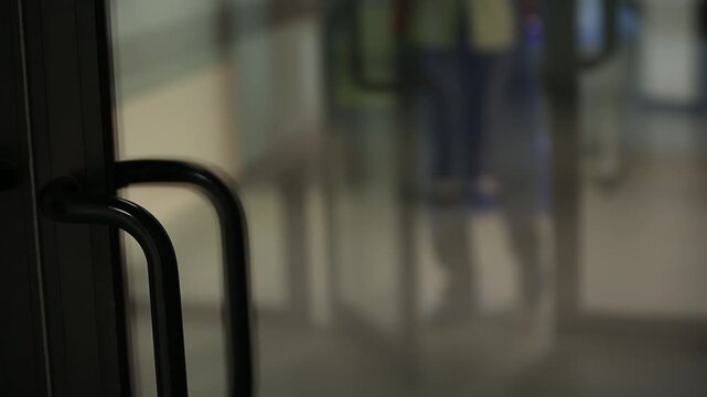 A person in a blue medical gown and white cuffs opens a set of glass doors with black handles, revealing a blurry hospital hallway.