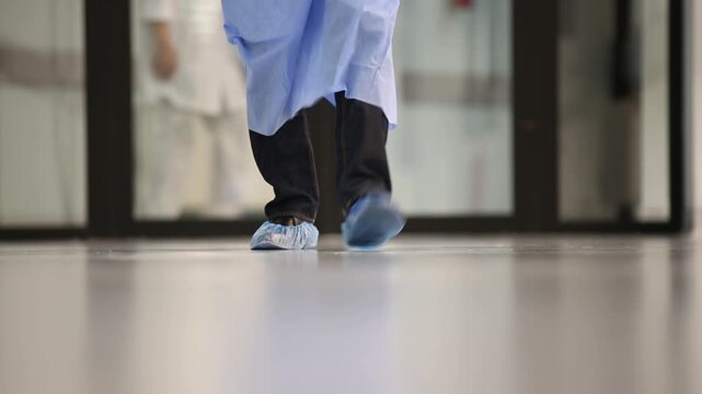 A person in a blue medical gown and white cuffs opens a set of glass doors with black handles, revealing a blurry hospital hallway.
