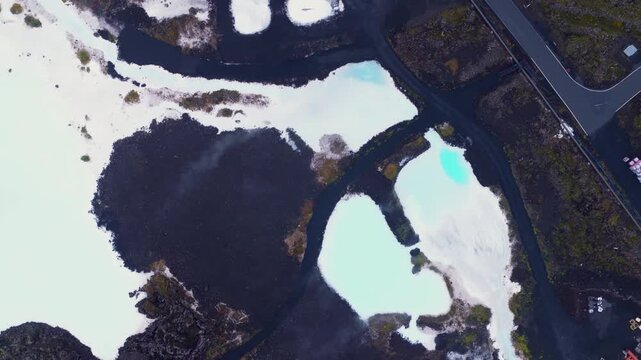 Smooth drone flight over Iceland's Blue Lagoon, showcasing milky blue geothermal pools, dark volcanic rock, and a road on the right side.
