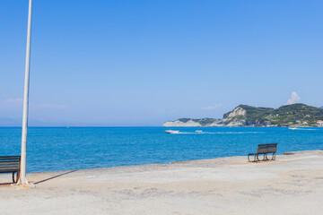 Solitary bench on concrete promenade facing crystal clear turquoise Mediterranean waters with distant islands under perfect blue sky.