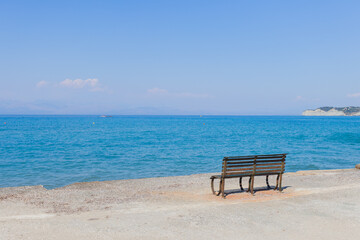 Solitary bench on concrete promenade facing crystal clear turquoise Mediterranean waters with distant islands under perfect blue sky.