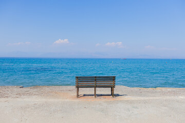 Solitary bench on concrete promenade facing crystal clear turquoise Mediterranean waters with distant islands under perfect blue sky.