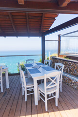 Oceanfront restaurant terrace with white wooden tables under pergola and glass panels overlooking scenic coastal landscape in bright sunlight.