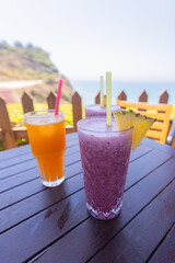 Two vibrant drinks — a purple pineapple smoothie and a bright orange juice — served on a rustic wooden table with an ocean cliff view, perfect for themes of travel, leisure, and coastal dining.