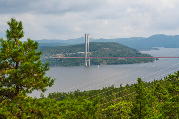 Fototapeta premium High Coast Bridge, a cable stayed structure in Sweden, spans two islands over Angermanalven river beneath a cloudy sky, framed by pine trees and power lines