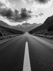 Naklejka premium A black and white shot of a long empty highway running through a desolate landscape, with majestic mountain peaks in the distance.