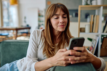 Smiling woman relaxing on a cozy sofa in her living room, happily using her smartphone for browsing and connecting online