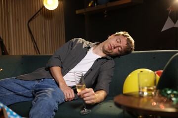 Man with glass of alcohol suffering from hangover on sofa in messy room after party