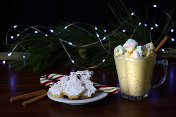Eggnog served in a glass, accompanied by a cinnamon stick and Christmas tree-shaped cookies coated...
