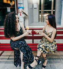 Two young friends, both in summer dresses and sunglasses, pose with ice cream in the street. One...