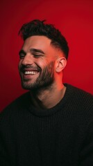 Obraz premium Smiling young Caucasian man with short dark hair and a beard, wearing a black sweater, against a red background.