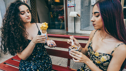 Two young friends, both in summer dresses and sunglasses, pose with ice cream in the street. One...