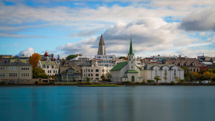 Reykjavík City skyline over Lake Tjörnin, with Fríkirkjan in the foreground, traditional houses lining the shore, and Hallgrimskirkja rising behind, a tranquil blend of heritage and modernity.