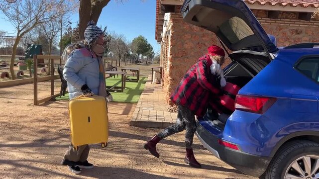 Two female friends loading suitcases into the trunk of a blue car, preparing to leave a rustic countryside cabin after a winter weekend trip