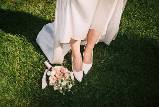 Bride in white heels with bouquet on grass
