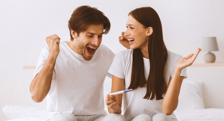 Happy excited emotional young caucasian man and woman hold pregnancy test in bedroom interior and...