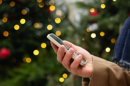 Man hand with smartphone against Christmas tree light garland and fairy lights. Xmas holiday. Close up.