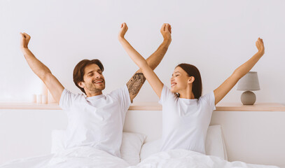 Happy attractive millennial european husband and wife stretching body and enjoying good morning in bed in bedroom interior, panorama. Serenity, energy, weekend and vacation, great mood and relaxation