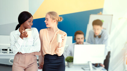 Diverse customer support operators on coffee break at modern call centre, space for text. Women telemarketing specialists with hot drinks making small talk at open space office
