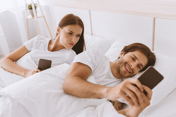 Fototapeta premium Offended angry young european wife looks at husbands phone and jealous, on bed in bedroom interior. Virtual cheating, gadget addiction during covid-19 lockdown. Chat in social networks and emotions