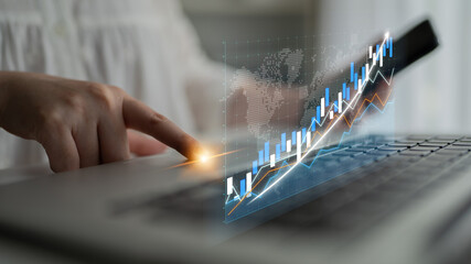 A businessperson uses a laptop to interact with a digital graph displaying upward trends. The image...