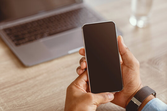 Unrecognizable millennial african american businessman holding smartphone with blank screen on workplace with pc in home office interior, close up. Website, offer and advertising, work and business - Powered by Adobe