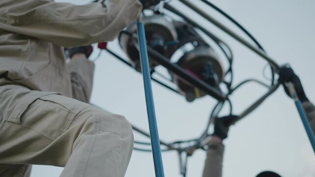 man assists woman with burner rig installation inside wicker basket during hot air balloon preflight setup by roadside under cloudy sky with red envelope and lush grassy field backdrop