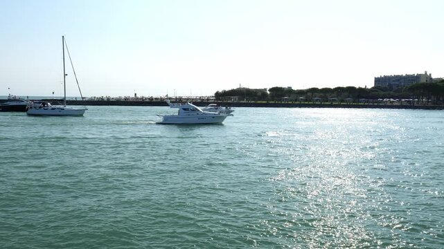Many large white yachts sail from the sea along the bay toward the city near the shore. Lots of people. Vacation in Italy. August 19, 2025. Caorle, Italy.
