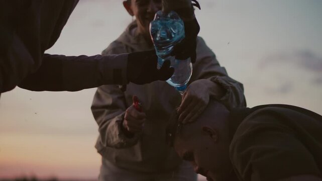 close up of woman holding lighter against man hair tip while water pours from bottle to quench flame in sunset field setting capturing tense instructive moment with protective intervention