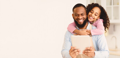 Free Time Together. Portrait of positive black man using digital tablet together with little daughter, smiling girl embracing dad from behind, odering groceries online or searching new recipe