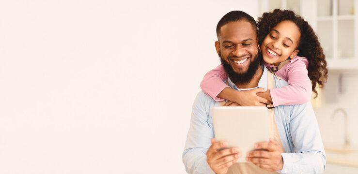 Free Time Together. Portrait of positive black man using digital tablet together with little daughter, smiling girl embracing dad from behind, odering groceries online or searching new recipe - Powered by Adobe