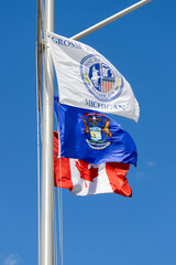 Three flags of Gross Ile township, Michigan state and Canada flag on single post against blue sky at USA Canada border..