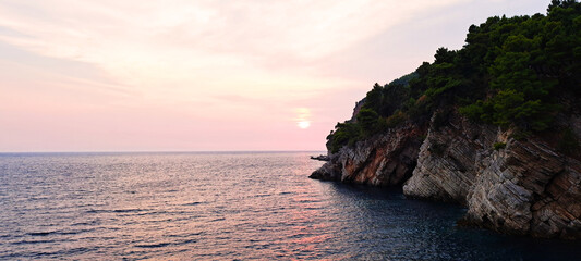 Dramatic rocky coastline with layered cliffs and lush green pines at sunset, overlooking calm blue sea waters reflecting soft pink and orange hues.
